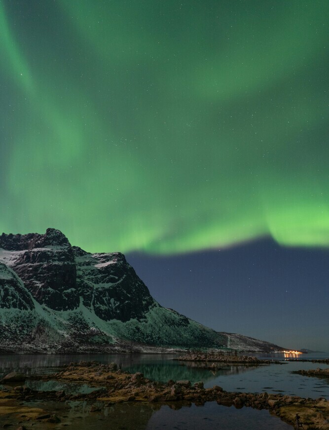 Colorful Northern Lights illuminating the night sky above mountains and reflecting over calm water below.