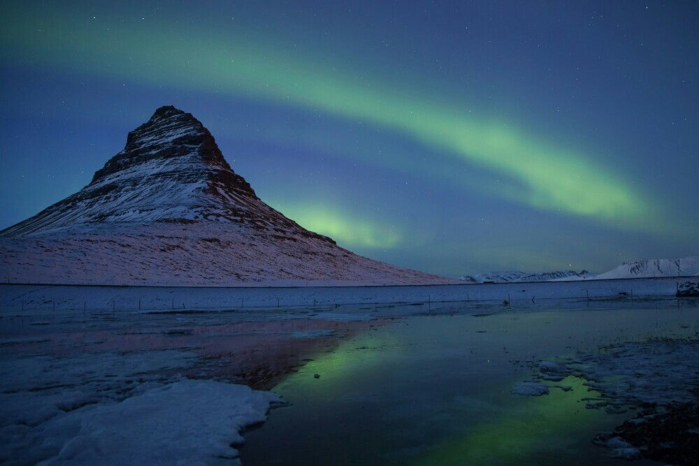 A stunning view of Kirkjufell mountain illuminated by the Northern Lights, with a cascading waterfall in the foreground under a starry night sky in Iceland