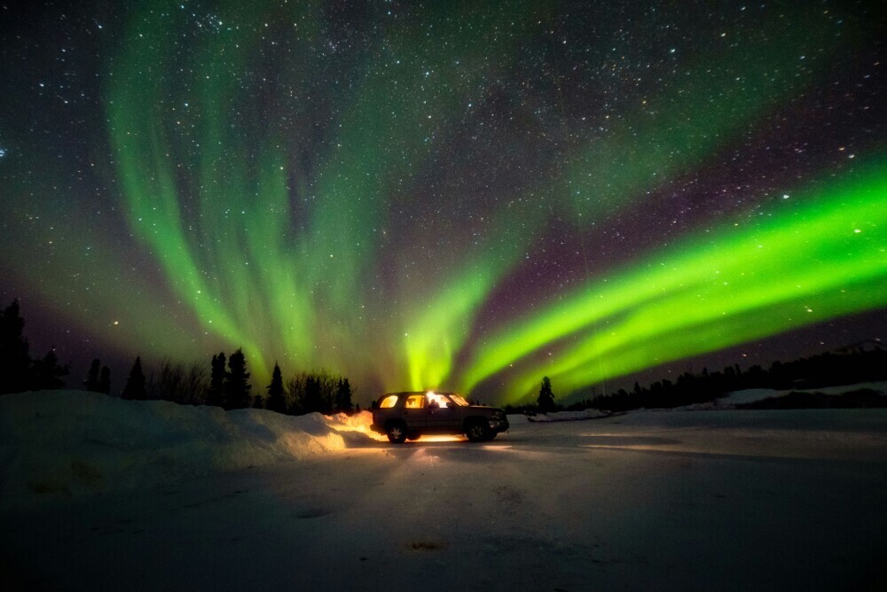 Car parked on snowy ground under vibrant green and purple Northern Lights in a clear night sky.