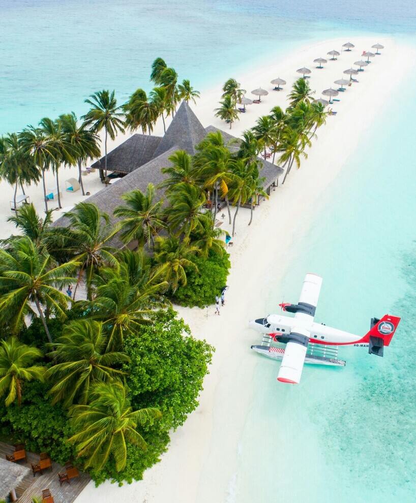 Aerial view of a pristine tropical island featuring lush green palm trees, a thatched-roof resort, and a turquoise lagoon. A seaplane is parked on the white sandy beach.