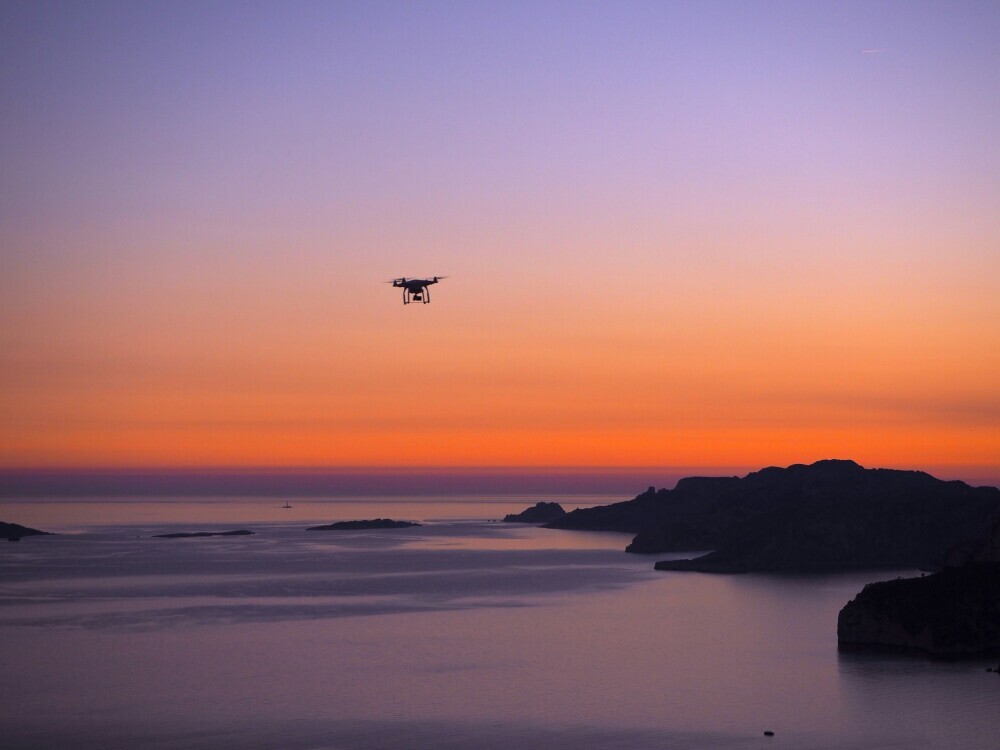 A drone flies high in a vibrant orange sky above snow-capped mountains and an expansive ocean.