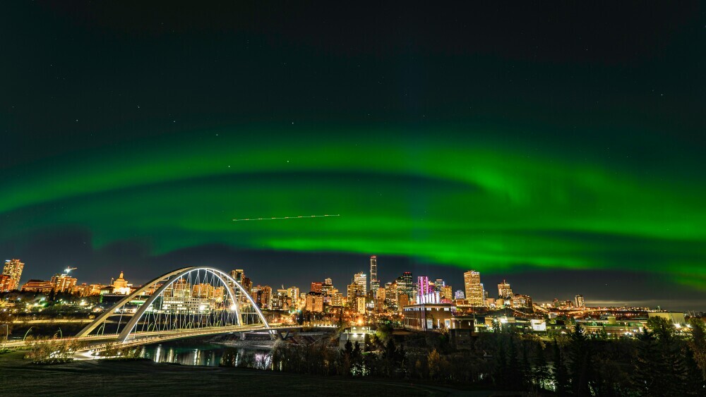 Northern Lights glowing vividly in green arcs above a brightly lit urban skyline and an illuminated bridge at night.