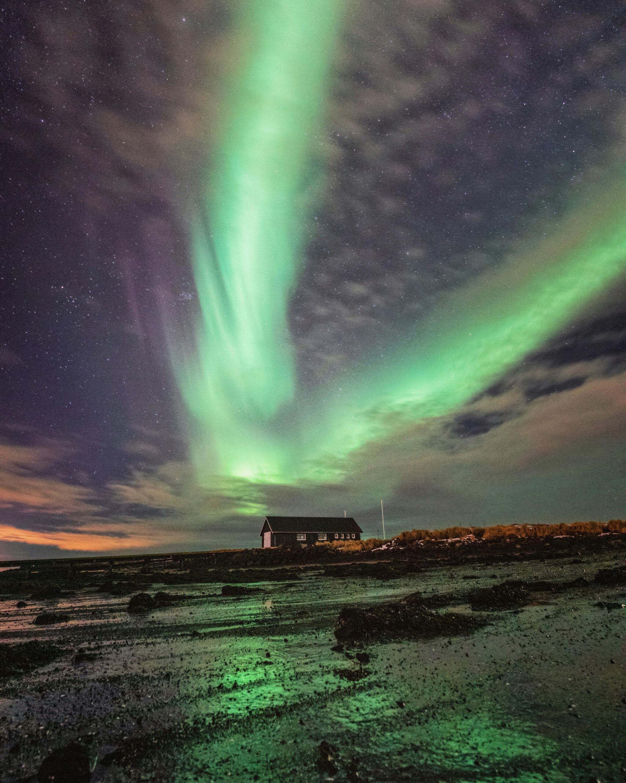 A small cabin nestled on a rocky beach, illuminated by a vibrant green Northern Lights display in the night sky.