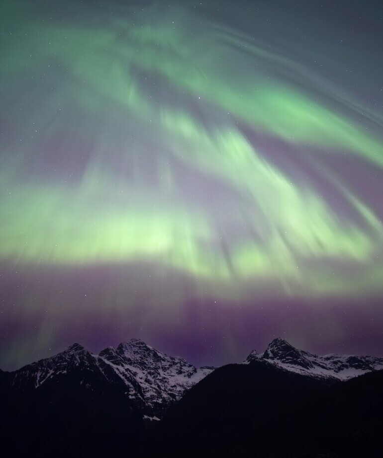 A mesmerizing display of the Northern Lights (Aurora Borealis) casting green and purple hues over snow-capped mountains under a starry night sky.