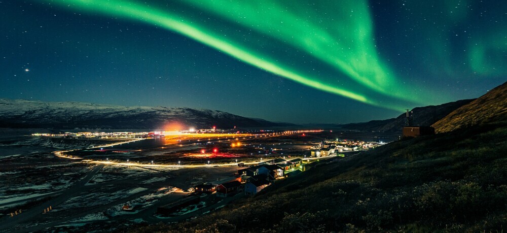 Stunning green Northern Lights illuminating the night sky above a vibrant cityscape, with colorful city lights reflecting off the surrounding majestic mountains.