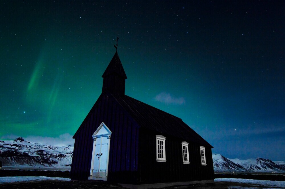 A dark wooden church under the glowing green Northern Lights in a snowy mountainous landscape.