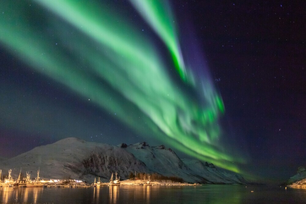 Northern Lights over a snowy mountain landscape with boats docked in a harbor.