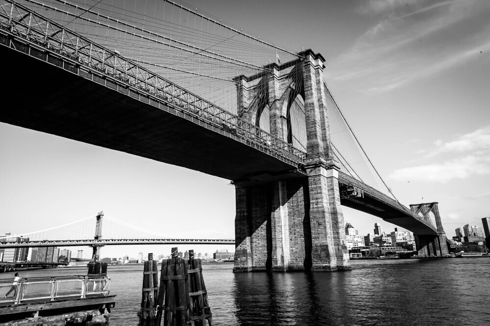 Black and white photograph of the Brooklyn Bridge with its intricate cables and towering stone pillars, captured from a low angle with the Manhattan Bridge in the background.