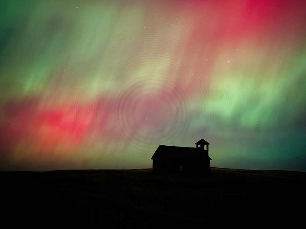 A vibrant display of red and green Northern Lights illuminates the night sky above a silhouetted rural building.